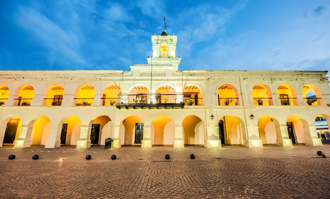 A historic colonial-style building with arched doorways and balconies, warmly lit at dusk, stands on a cobblestone plaza under a blue sky—capturing the essence of Northern Argentina and its architectural wonders.
