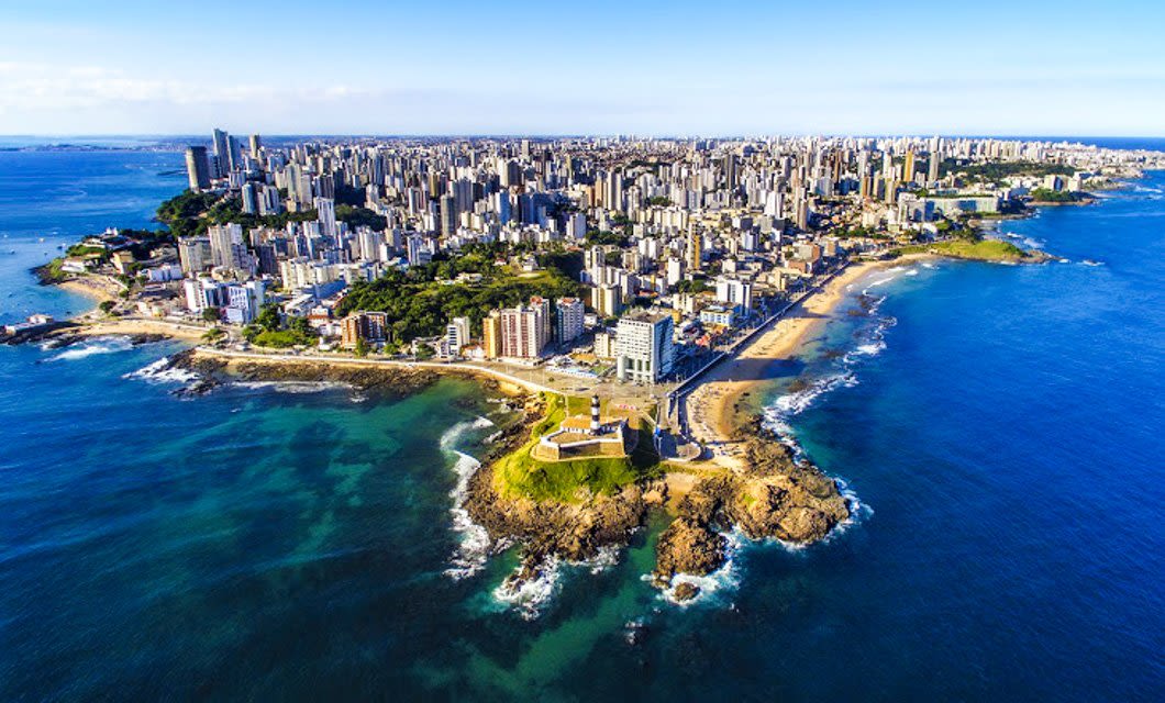 Aerial view of a coastal city in South America with many tall buildings, sandy beaches, green areas, and a prominent peninsula surrounded by blue ocean waters—perfect for a Deluxe Tour under a clear sky.