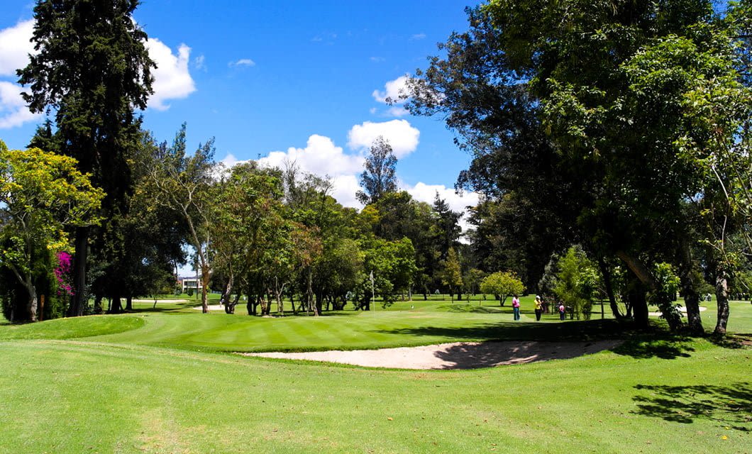 A sunny golf course with lush green grass, trees surrounding the fairway, a sand bunker in the foreground, and several people walking in the distance—perfect for a South America Golf Tour under Brazil’s bright blue sky.