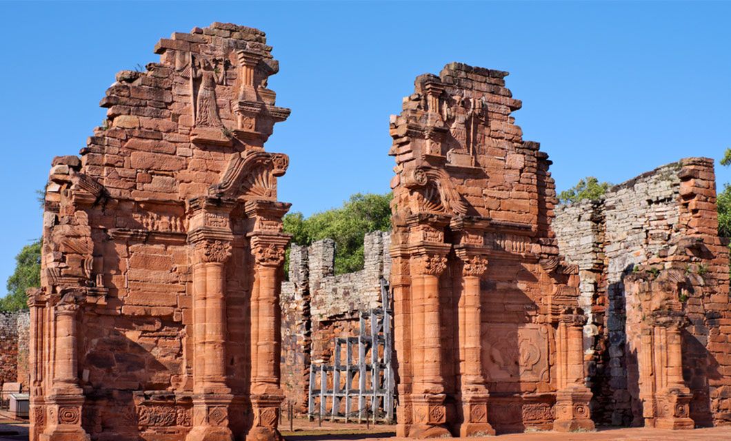 Ancient red stone ruins with tall columns and ornate carvings, partially crumbled, stand against a clear blue sky and lush greenery—a highlight on the Coast to Coast Tour.