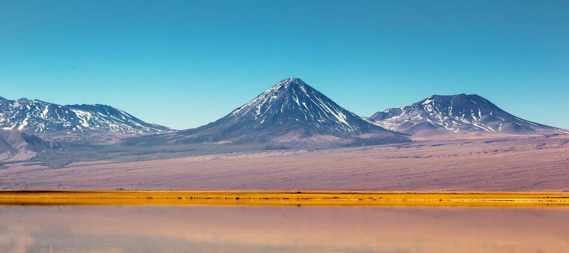 Snow-capped mountains with a sharp peak rise above a barren, sandy desert landscape, reflected in a calm body of water under a clear blue sky—a breathtaking view often featured on Bolivia and Chile tours.