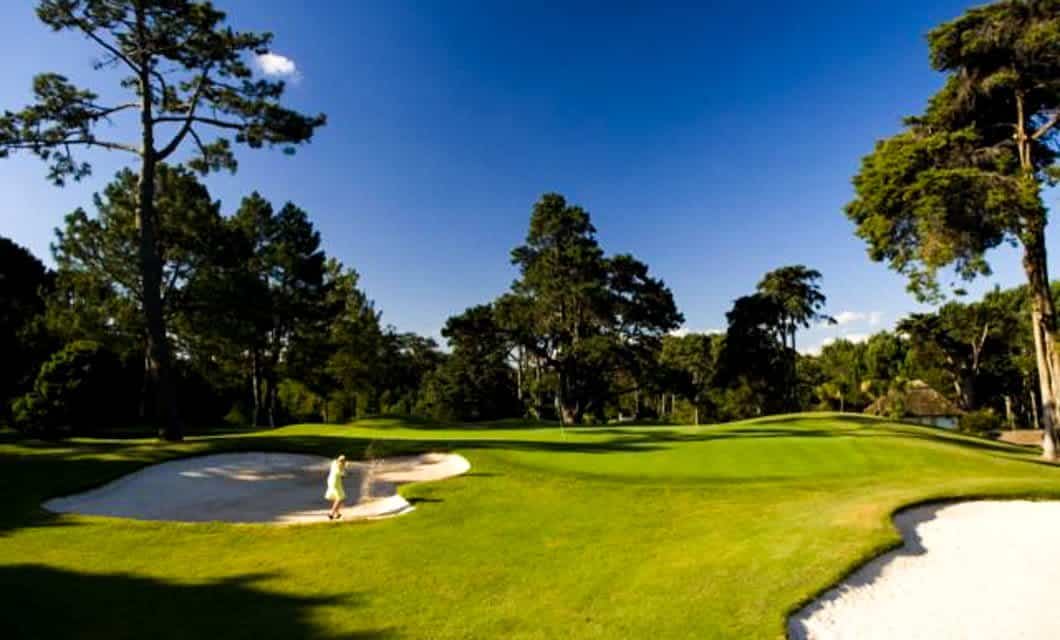 A person stands in a sand bunker on a lush green Argentina golf course surrounded by tall trees under a clear blue sky. Another sand bunker is in the foreground.
