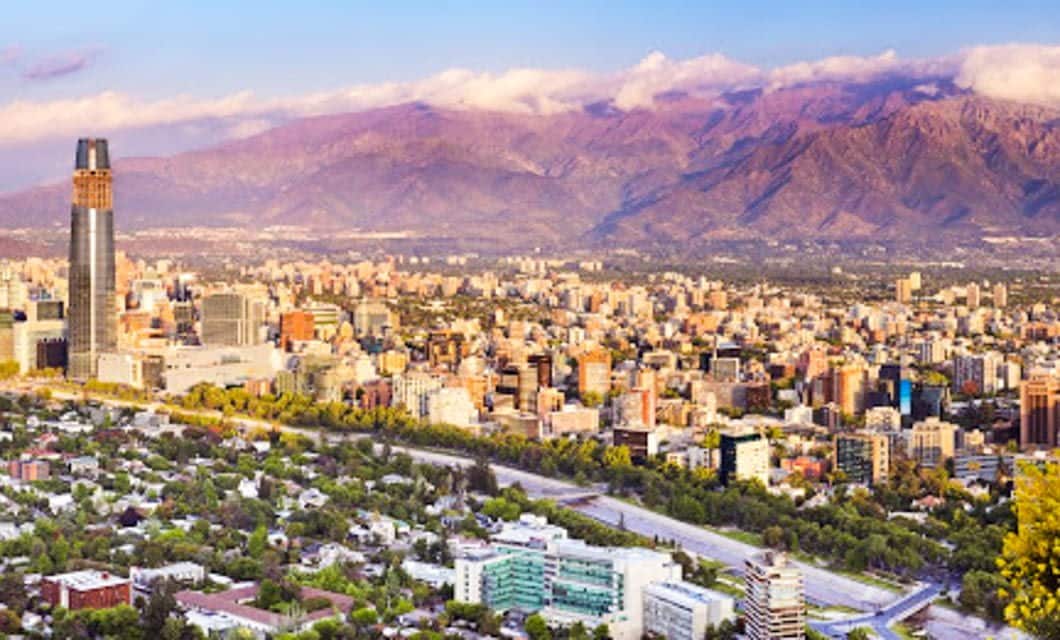 A vibrant cityscape in Chile with tall buildings and a mix of modern and older architecture, set against purple mountains under a partly cloudy sky. Green trees and streets are visible—an ideal setting for an unforgettable Eclipse Tour.
