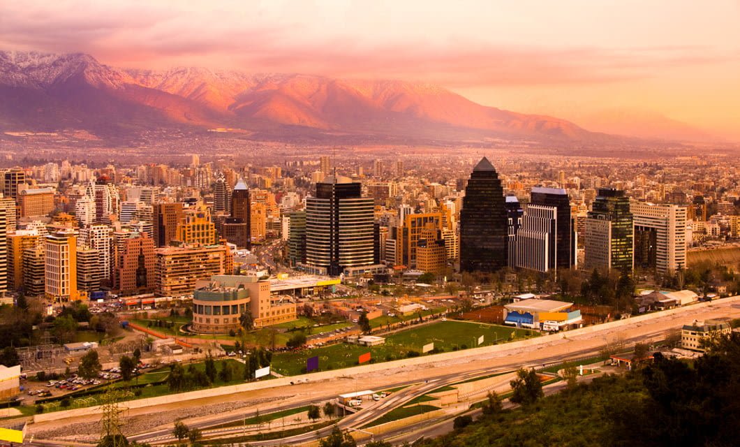 A panoramic view of a modern city like Buenos Aires, with tall buildings, green spaces, and mountains in the background, all bathed in warm, golden sunset light.
