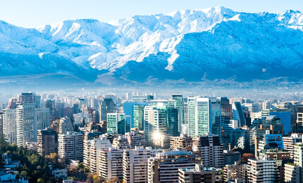 Modern cityscape with tall buildings in the foreground and towering snow-capped mountains in the background under a clear blue sky, evoking the charm of Buenos Aires or a view you might enjoy on a Santiago Wine Tour.
