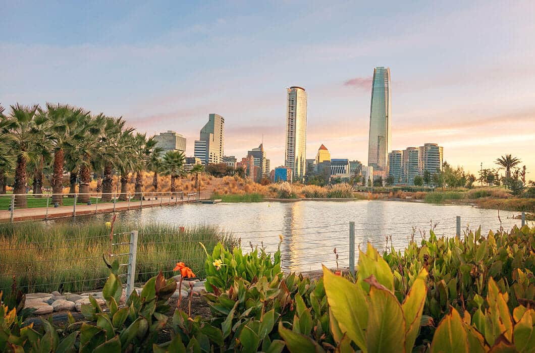 A view of a modern city skyline with tall buildings behind a park featuring a pond, green plants, and palm trees under a pastel evening sky, nestled near tranquil lakes.