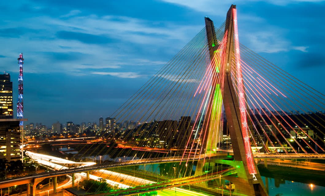 Cable-stayed bridge illuminated with colorful lights at dusk, spanning over a river in a vibrant South American cityscape with modern buildings and traffic trails—perfect for a Deluxe Tour experience.