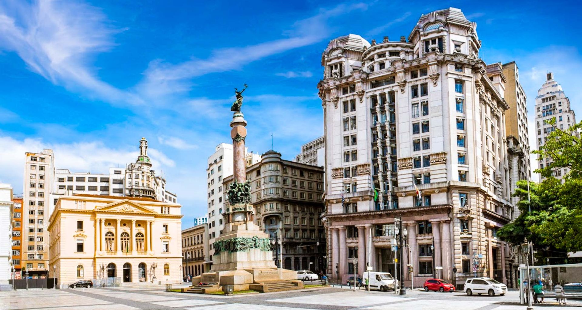 A sunny city square with a tall monument in the center, where locals sometimes dance tango, surrounded by grand historic buildings, including a yellow neoclassical structure and ornate white high-rises under a bright blue sky.