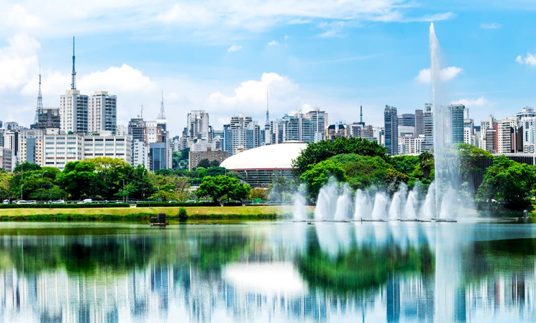 A cityscape with tall modern buildings, a large domed structure, and water fountains in a lake, surrounded by trees under a bright blue sky—once part of the historic Brazil Gold Route, this scene blends modern life with storied heritage.