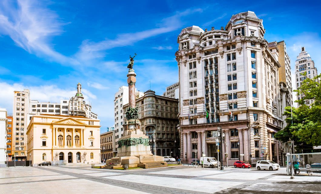 A sunny city square with historic buildings, a yellow theater, a tall ornate monument with statues, and a large white classical building under a bright blue sky. The lively scene hints at the passionate spirit of tango as cars and people bustle in the background.