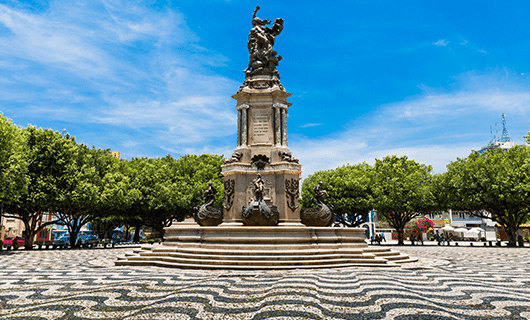 A tall monument with ornate statues stands in the center of a patterned cobblestone plaza in Rio de Janeiro, surrounded by green trees under a bright blue sky.