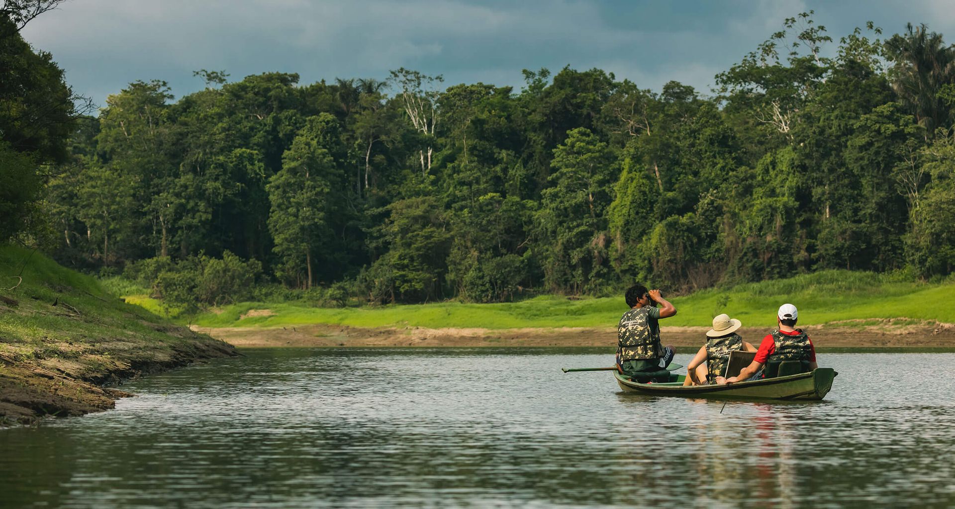 Three people in a small boat paddle along a calm river surrounded by dense, green jungle under a cloudy sky, immersing themselves in a Remote Amazon Tour to explore and observe the untouched beauty of nature.