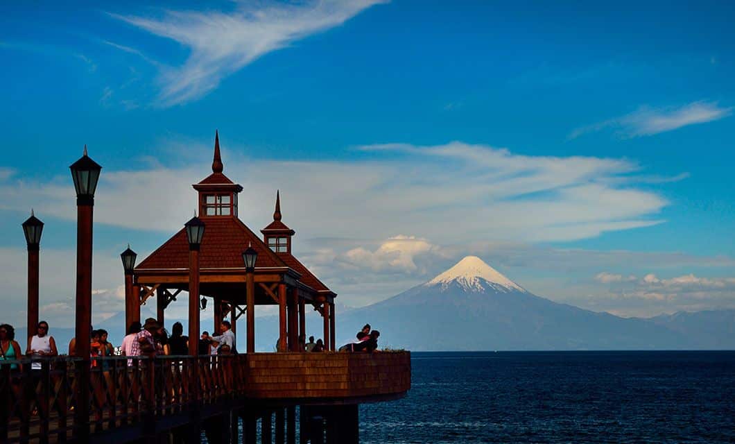 A wooden pier with gazebos and people overlooks a large blue lake near Bariloche. In the distance, a snow-capped volcano rises against a bright sky with scattered clouds, evoking the beauty of a classic Patagonia Tour.