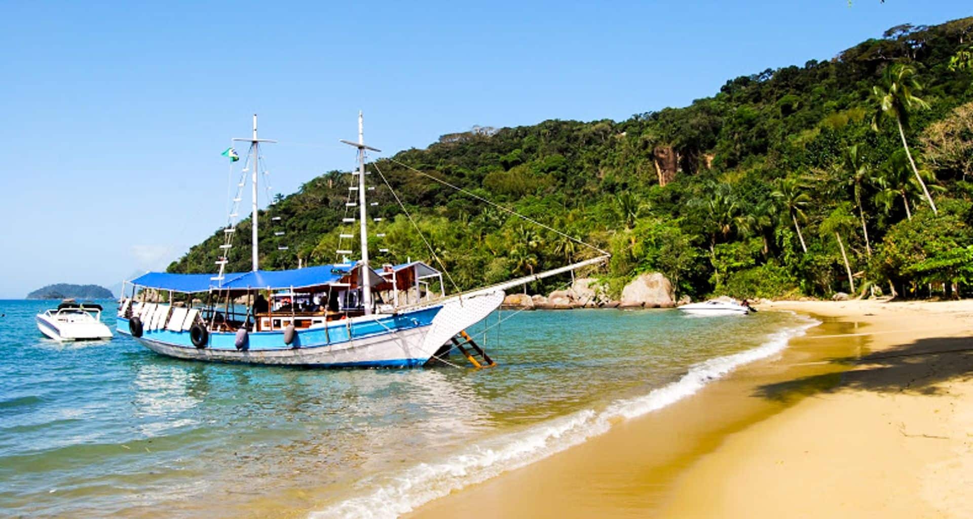 A white and blue sailboat is anchored near the golden sandy shore of a tropical island in Paraty, with lush green palm trees and forested hills in the background under a clear blue sky.