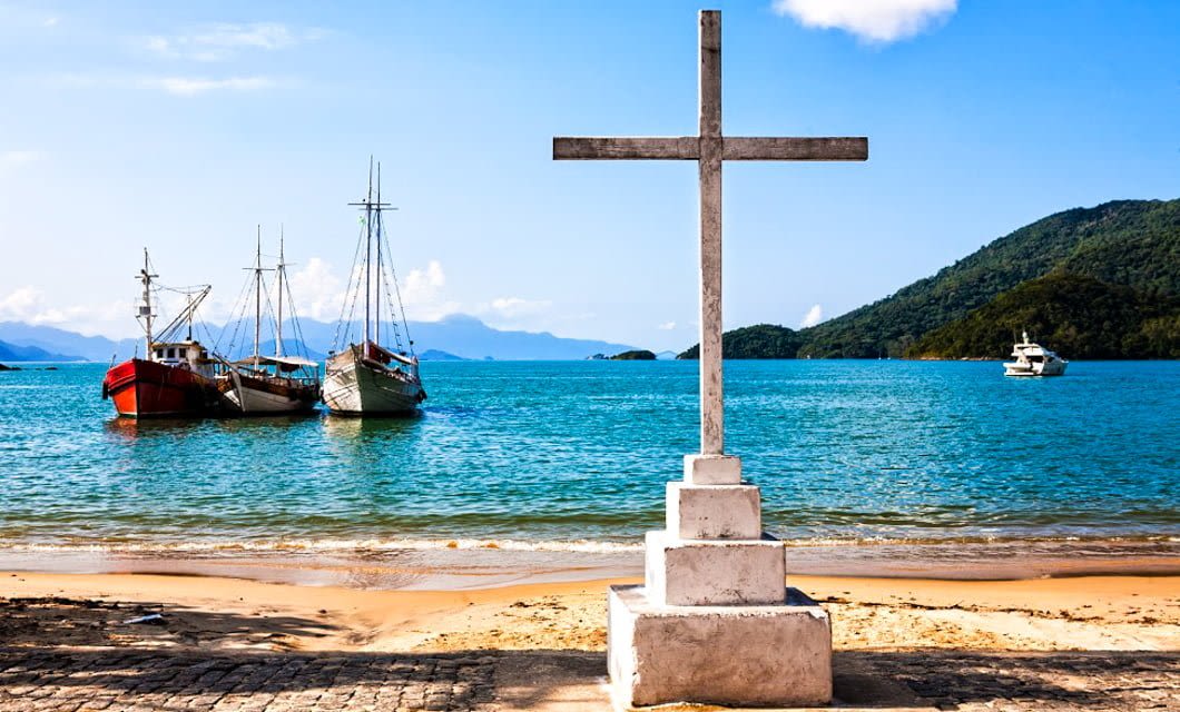 A stone cross stands on a beach in Paraty, overlooking calm blue water with three boats anchored near the shore and green hills in the background under a clear tropical island sky.