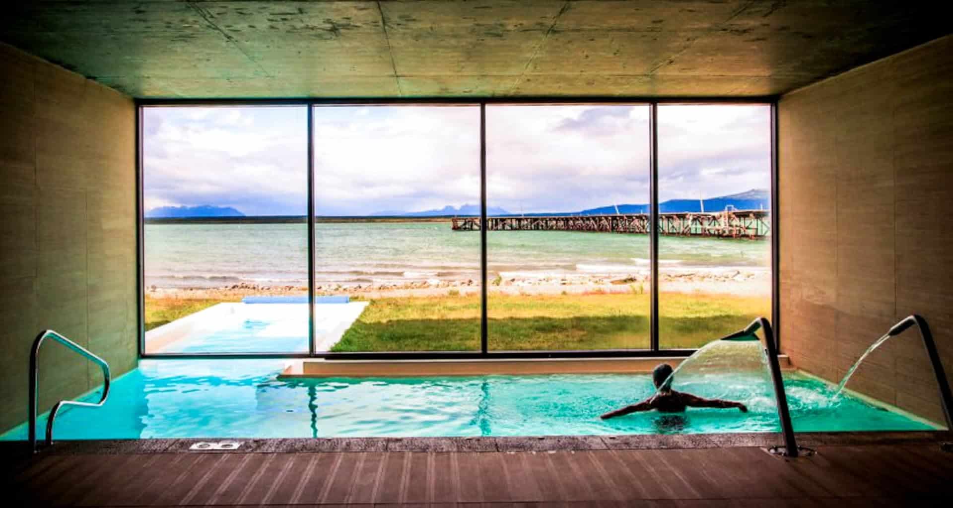 A person relaxes in an indoor pool with water jets at the luxury hotel Singular Patagonia, facing large windows that reveal a grassy area, a pier, and distant mountains under a cloudy sky.