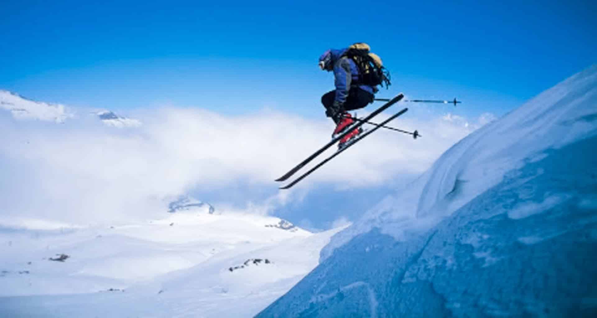 A skier wearing winter gear and a backpack is mid-air while skiing in Ushuaia, leaping off a snowy slope surrounded by majestic, snow-covered mountains and a bright blue sky.