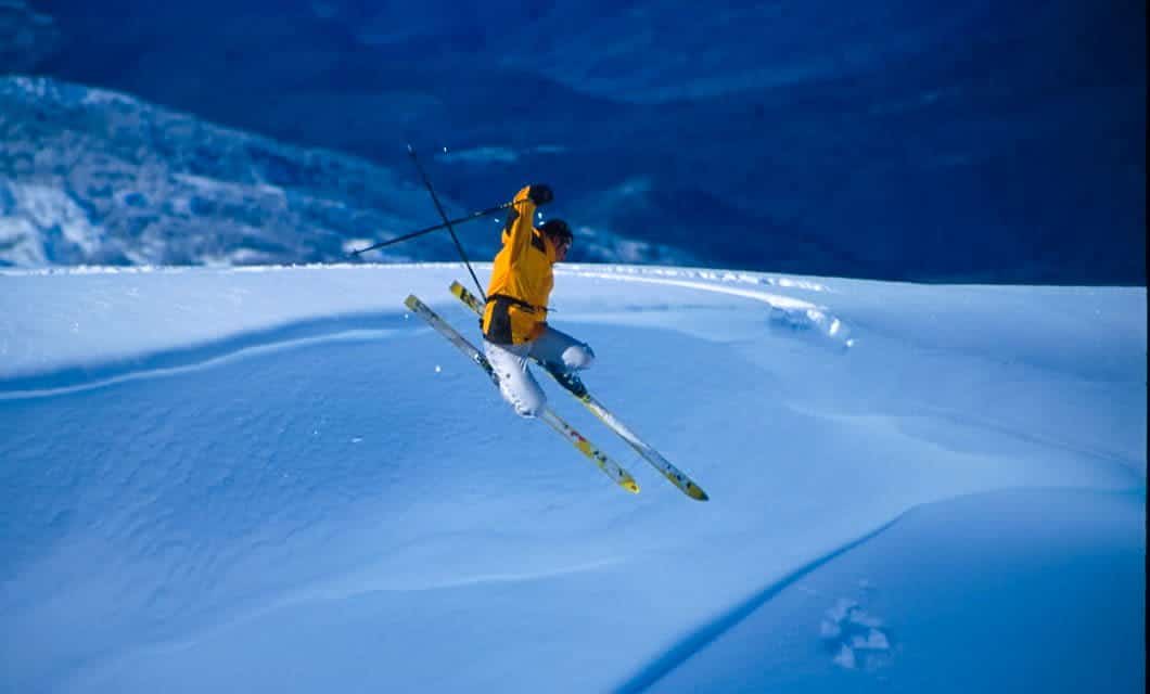 A skier in a yellow jacket and black pants jumps off a snow-covered slope, holding ski poles in both hands, with snowy mountains and a blue sky in the background—capturing the thrill of skiing in Ushuaia.