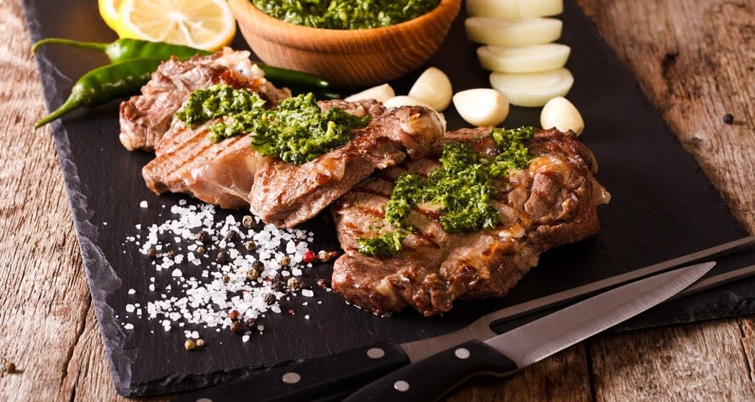 Three grilled steaks topped with green chimichurri sauce are served on a black slate platter with lemon slices, garlic cloves, sliced onions, and green chili—a feast inspired by Argentina’s rich food and wine tour traditions.