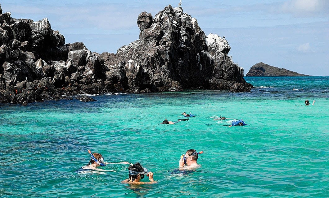 People snorkeling in clear turquoise water near rugged, rocky cliffs under a partly cloudy sky. This Galapagos tour scene offers a multisport adventure, with swimmers exploring the vibrant sea along the unique subtropical coastline.
