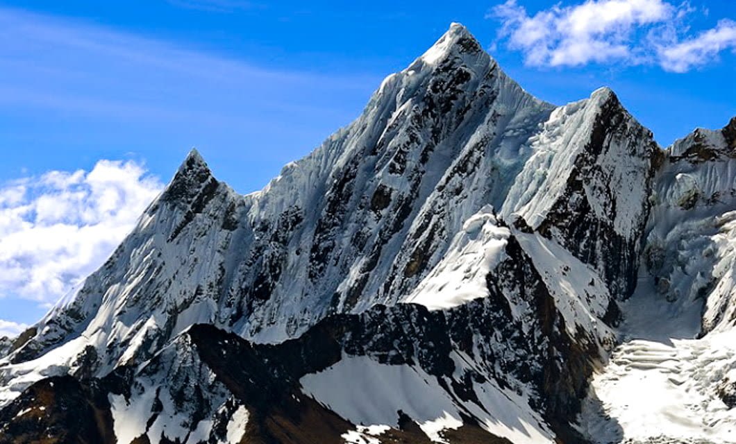 Snow-covered jagged mountain peaks of the Cordillera Blanca rise sharply against a bright blue sky with scattered clouds. Sunlight highlights the crisp edges and rugged slopes, capturing the dramatic beauty near Huaraz on a classic Peru trek.