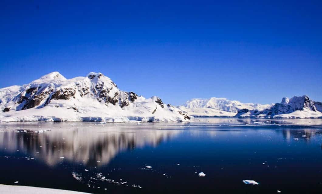 Snow-covered mountains reflected in calm, dark blue water under a clear sky capture the tranquil beauty witnessed on an Antarctic expedition, with small chunks of ice floating peacefully on the surface.