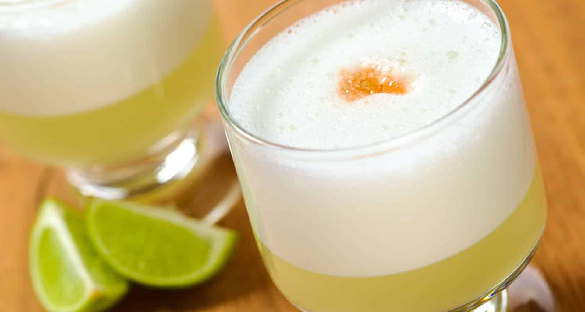 Two glasses of frothy yellow cocktail, likely pisco sours, with a drop of bitters on top. Lime wedges rest beside the drinks on a wooden surface—perfect for savoring during a Peru winery gastronomy tour.