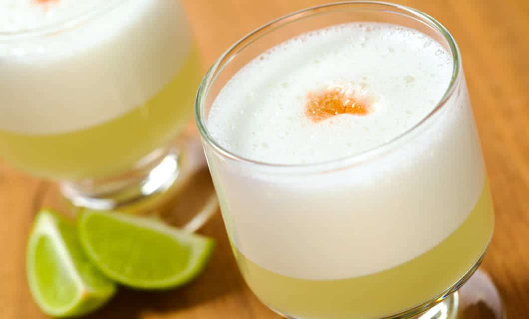 A glass of frothy yellow cocktail, garnished with a drop of bitters, sits on a wooden surface with lime wedges in the background—a perfect sip after a Peru tour or gastronomy tour exploring the country’s vibrant culinary scene.
