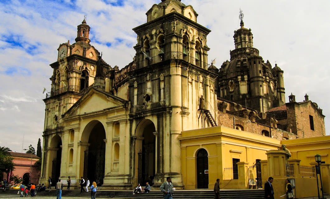 A historic baroque-style Jesuit cathedral with two tall towers and detailed architecture stands in a plaza, often called the intellectual heart of South America, with people walking and sitting nearby under a partly cloudy sky.