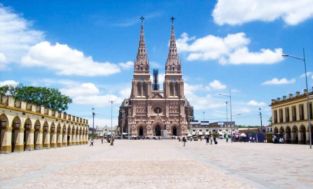 A large cobblestone plaza leads to a grand cathedral with two tall, ornate spires under a bright blue sky in Chile. Arched yellow buildings line the sides, evoking the charm often seen on a Patagonia railway tour through Argentina and Chile.