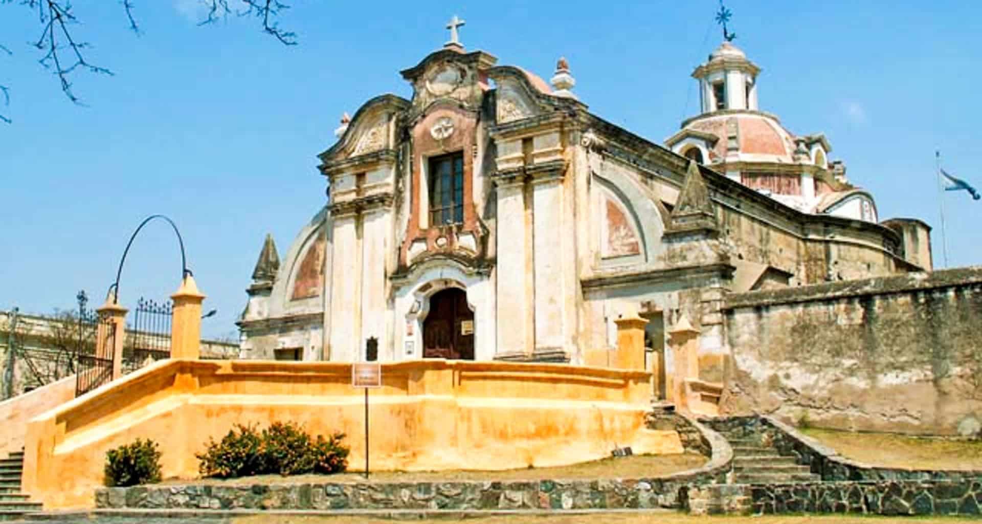 Historic Jesuit colonial-style church with weathered facade, ornate details, and domed roof stands as the intellectual heart of South America, set behind a yellow-orange wall and stairs amid stone pavement and greenery under a clear blue sky.