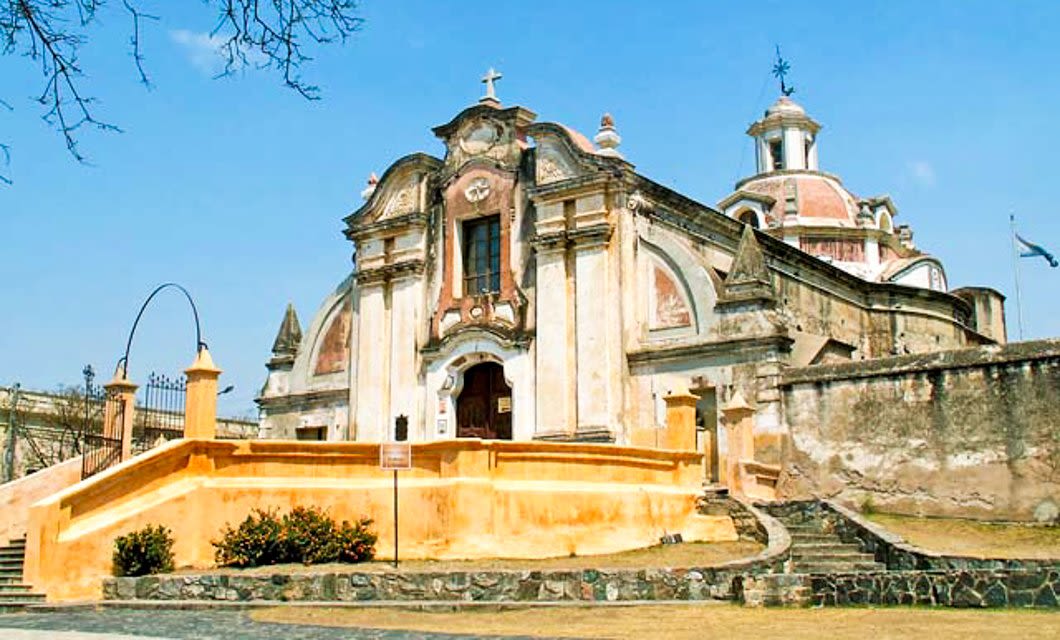 Historic baroque-style Jesuit church with weathered stone walls, arched entrance, and a dome, set against a blue sky. This landmark sits atop a raised platform with stairs, surrounded by stone walls and shrubs in the intellectual heart of South America.