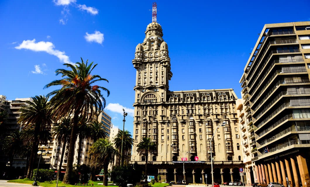 Historic Palacio Salvo stands tall under a bright blue sky, surrounded by palm trees and modern buildings in the heart of Montevideo, Uruguay.