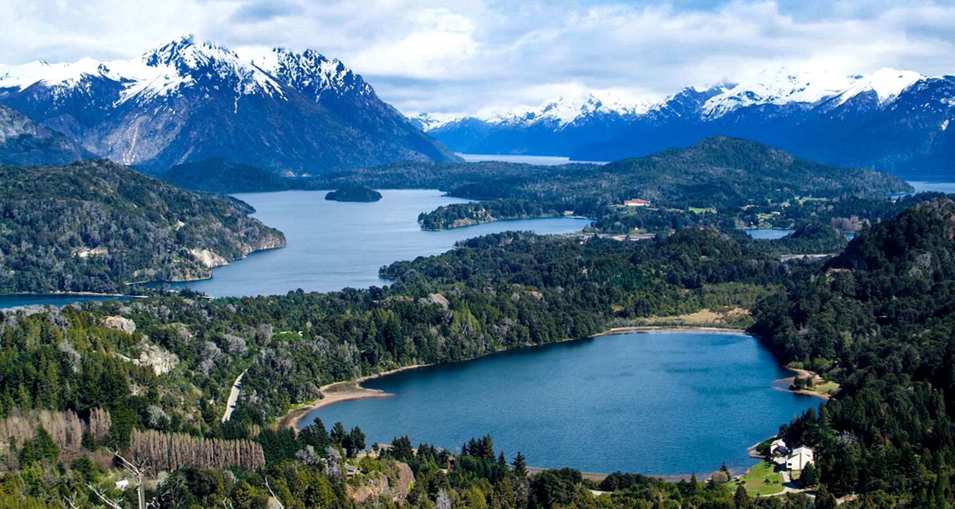 A panoramic view of a lush green landscape with sparkling lakes, cascading waterfalls, dense forests, and snow-capped mountains under a partly cloudy sky.