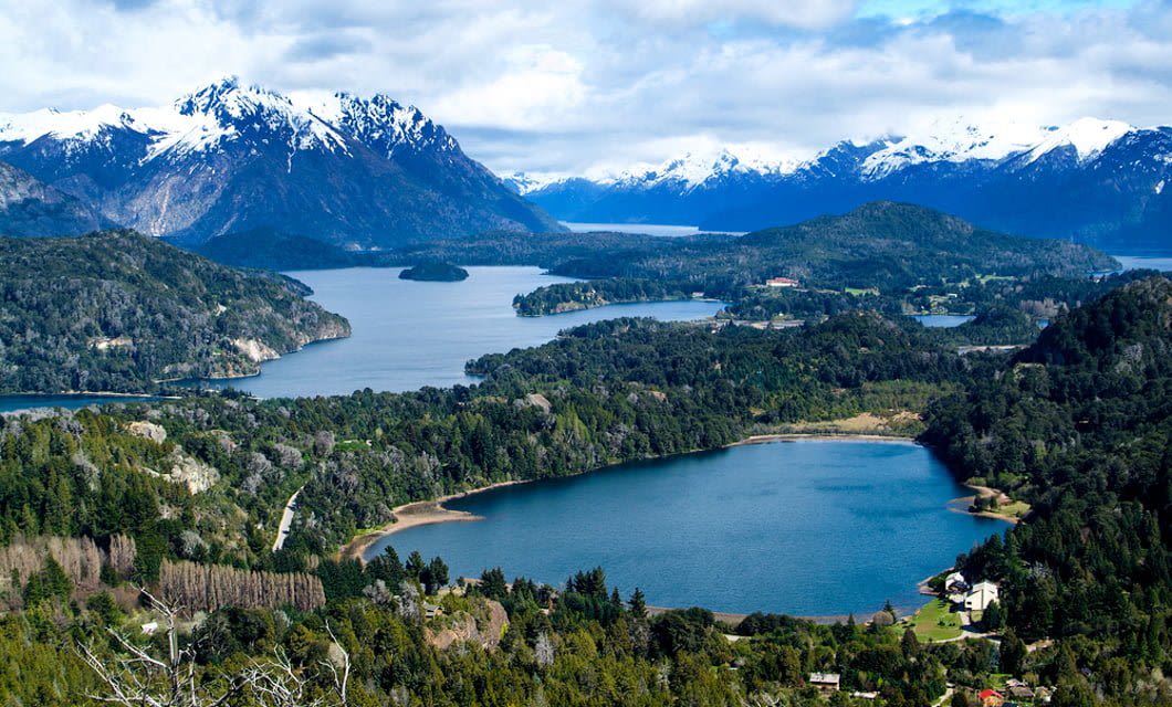A panoramic view of Patagonia Lakes with a lush green landscape, a large lake surrounded by forested hills, snow-capped mountains in the background, and scattered buildings near the shore under a partly cloudy sky.