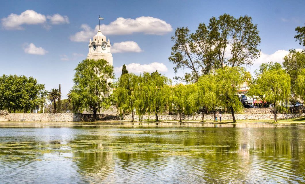 A white Jesuit clock tower rises above trees and a stone wall on the far side of a calm pond, with blue sky and scattered clouds in the background, evoking the Intellectual Heart of South America.