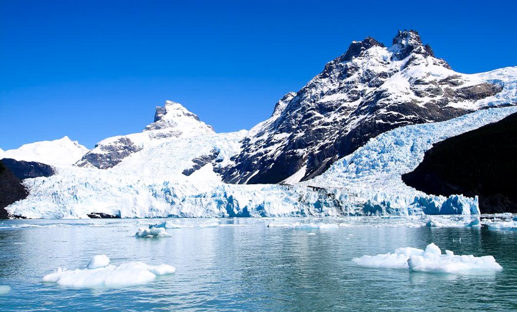 Snow-capped mountains and a glacier rise above a clear, icy lake with floating icebergs under a bright blue sky—an unforgettable sight on any Argentina travel adventure.