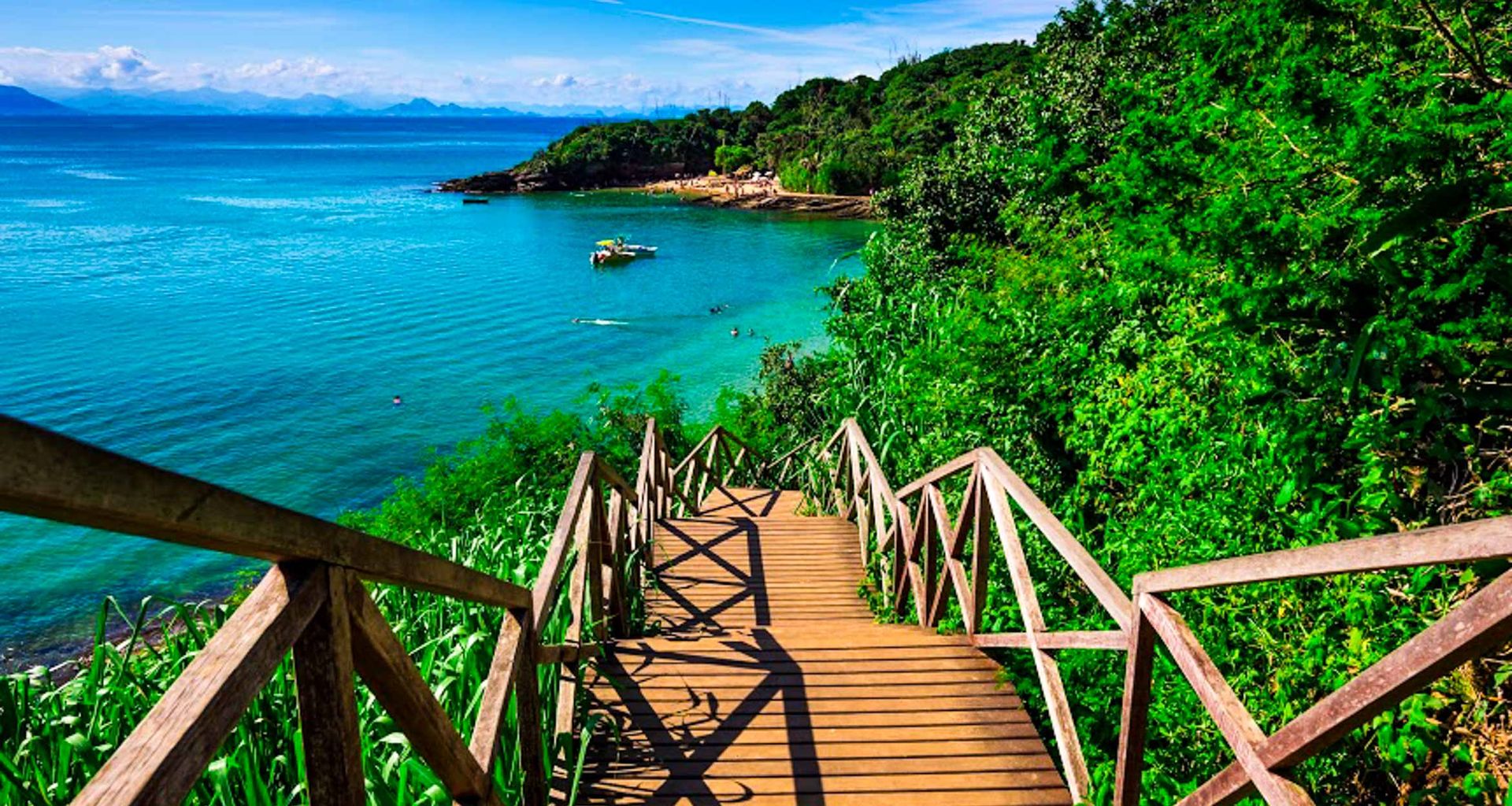 Wooden stairs surrounded by lush green plants lead down to a clear turquoise sea in Buzios Beaches, with a boat floating near the shore and distant mountains visible under the Rio de Janeiro blue sky.
