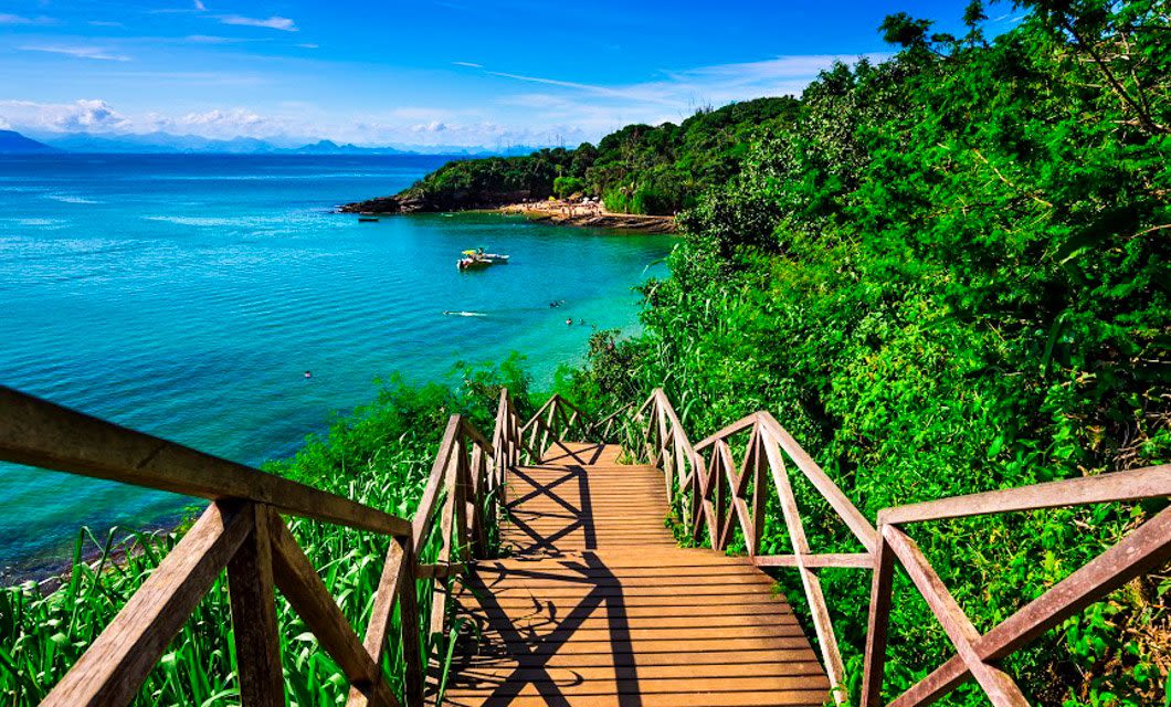 A wooden staircase descends through lush green vegetation toward a turquoise sea, reminiscent of Buzios Beaches, with boats floating on the water and distant hills under a bright blue sky.