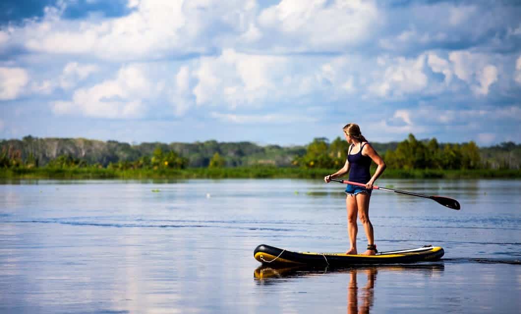 A person in a tank top and shorts stands on a paddleboard, paddling on a calm lake surrounded by greenery—reminiscent of the serene scenes witnessed during an Amazon Delfin II Cruise under blue skies with scattered clouds.