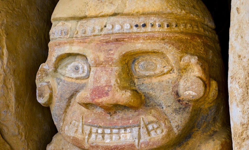 Close-up of an ancient carved stone face with large, wide eyes, a broad nose, and a detailed mouth showing teeth—much like relics you might spot on a Colombia Coffee tour. The figure wears a decorated headband and displays weathered textures.