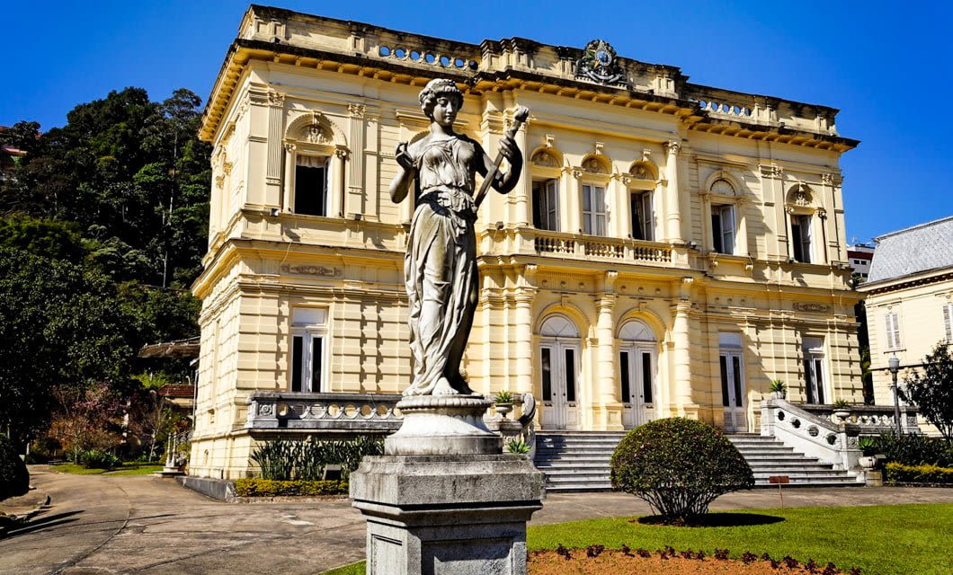 A classical stone statue of a woman stands before a grand, ornate yellow building along the Brazil Gold Route, with arched windows and lush greenery beneath a clear blue sky.