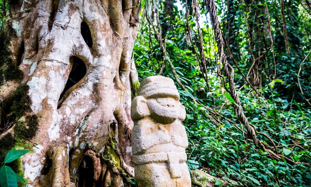 A stone statue with a rounded head and arms crossed stands beside a large, gnarled tree trunk in a lush, green jungle filled with vines and dense foliage—an ideal scene to discover on a Colombia Coffee Tour.