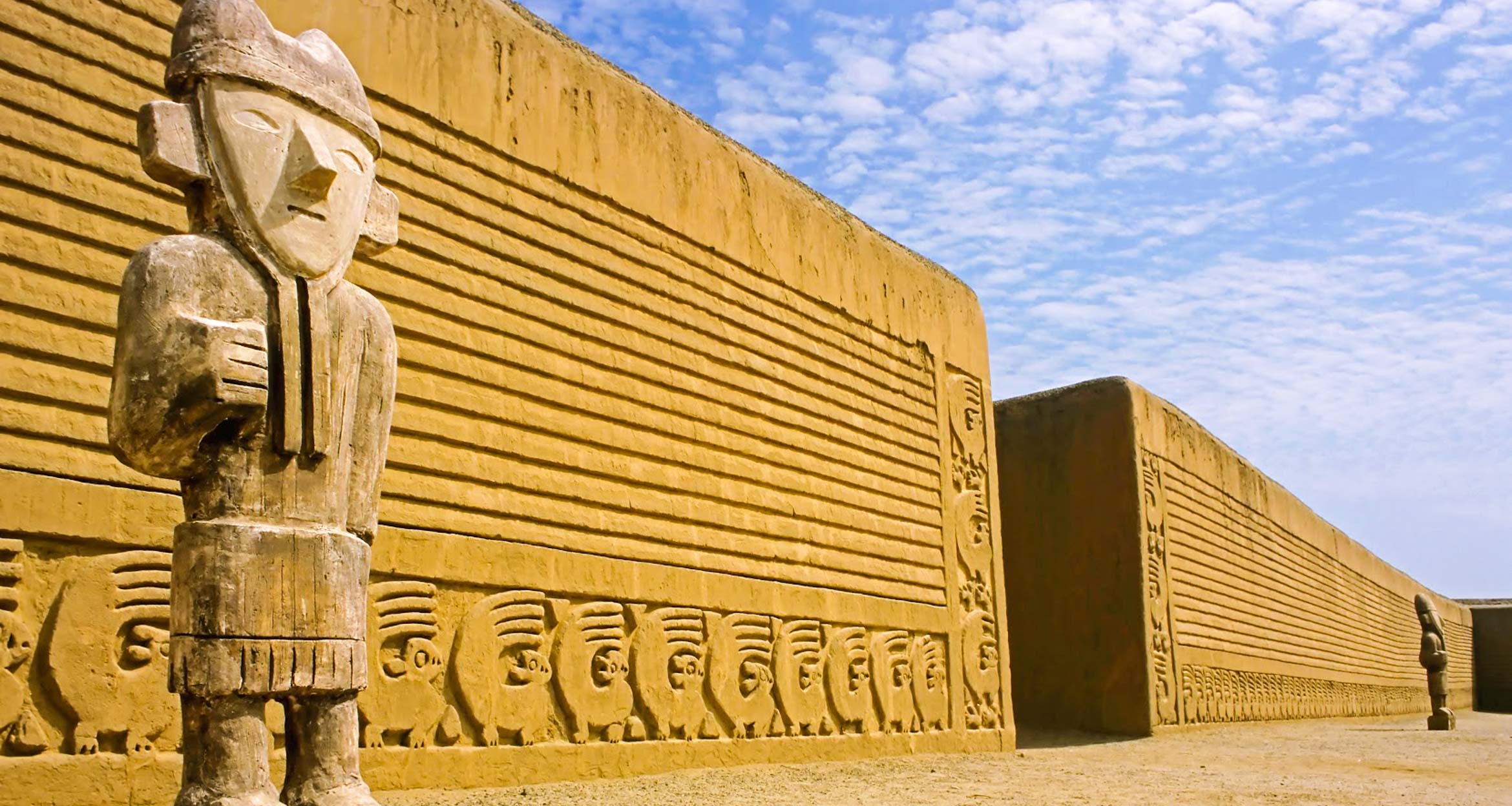 Large adobe walls decorated with intricate carvings and statues of ancient figures stand under a blue sky with scattered clouds, evoking the rich history explored on a Cajamarca Archaeology Tour or during visits to sites near Chiclayo and Trujillo.