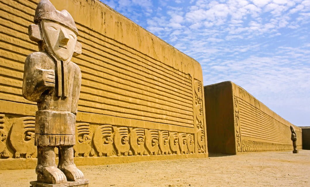 Ancient adobe statues stand beside a long, ornately carved wall under a blue sky at the archaeological site of Chan Chan, near Trujillo. The wall features intricate designs and geometric patterns, making it a highlight of any archaeology tour.