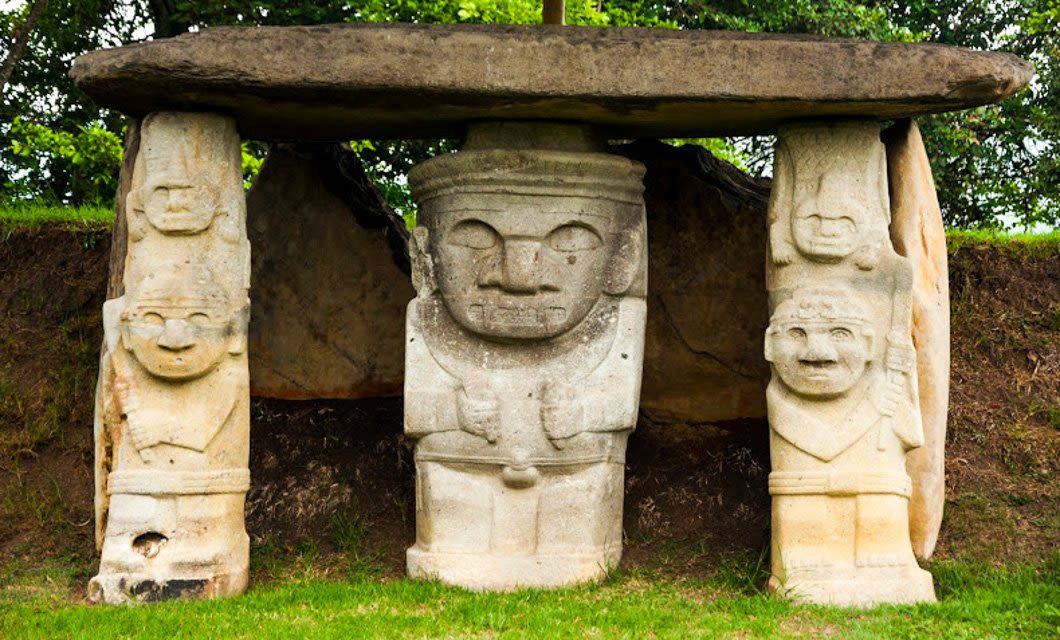 Three large stone statues with human-like faces and figures stand beneath a stone slab in the grassy San Agustín Ruins, surrounded by green trees and bushes—a highlight of any Colombia Archaeology Tour. The central figure is larger and more detailed than the others.
