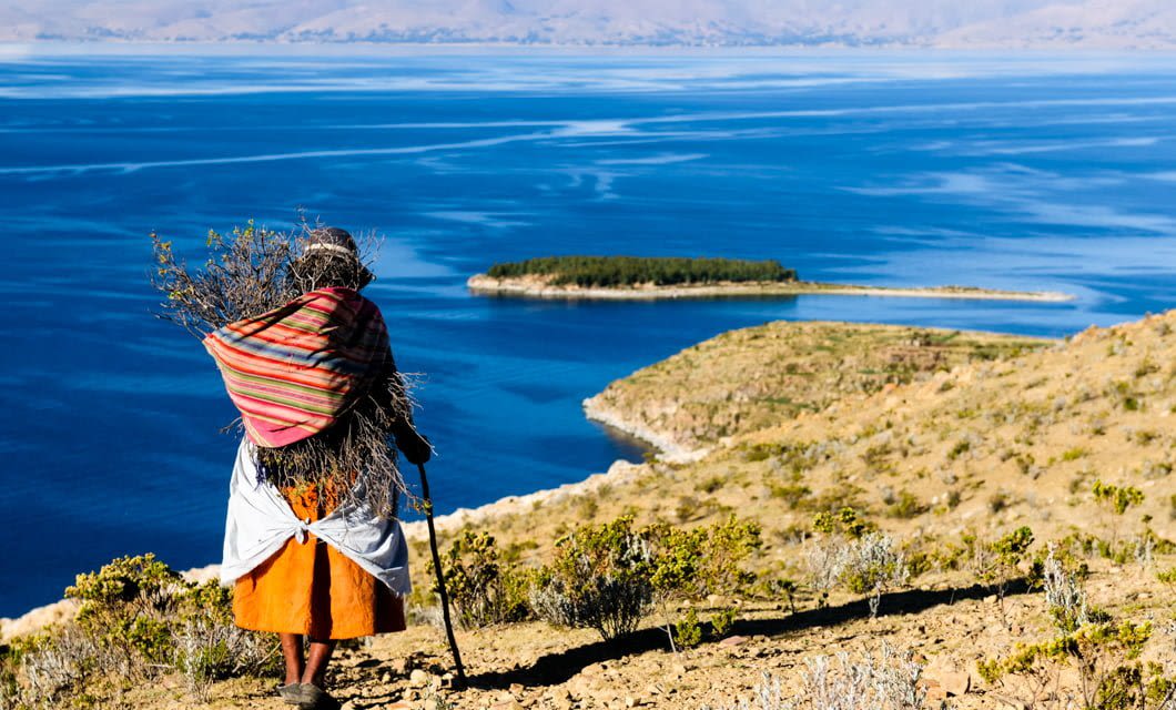 A person wearing colorful traditional clothing carries a bundle of branches on their back, walking with a stick toward a blue lake—perhaps a highlight on a Coast to Coast Tour—with an island and mountains visible in the background.