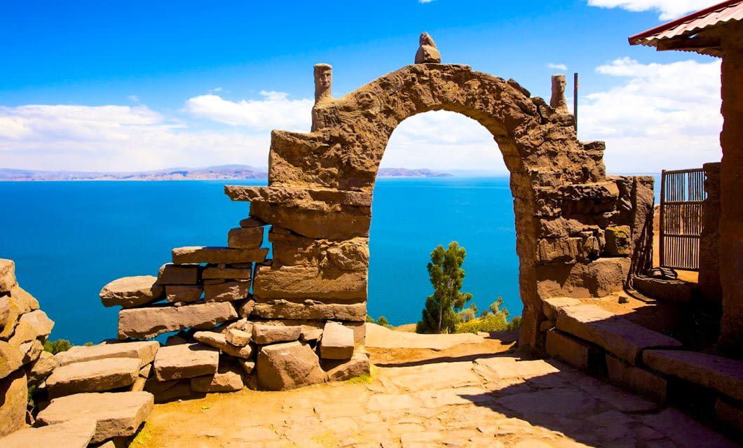 A stone archway with decorative carvings overlooks a deep blue lake and distant mountains under a bright, sunny sky. Some trees and scattered stone structures are visible near the arch, evoking the scenic beauty of Peru and Bolivia.