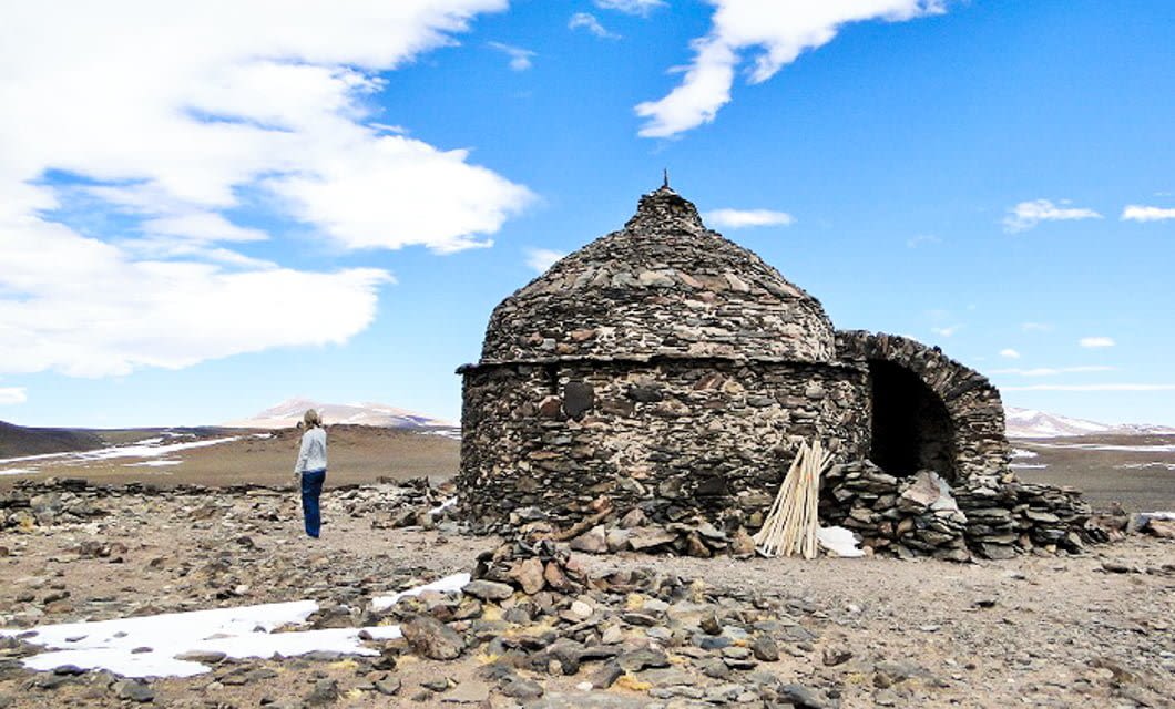A person stands near a round stone hut with a domed roof and arched entrance, set in the barren, rocky Andes landscape under a bright blue sky with scattered clouds—an image reminiscent of remote Velasco settlements.