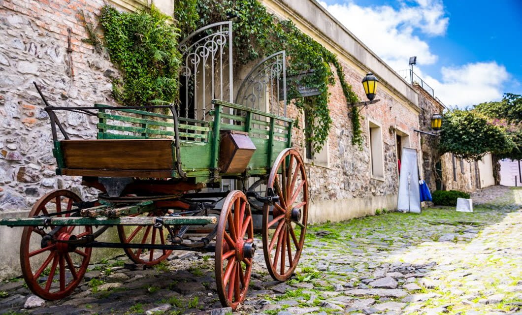 An old green wooden wagon with red wheels sits on a cobblestone street in Buenos Aires, beside a stone building covered in ivy, under a bright blue sky with scattered clouds.